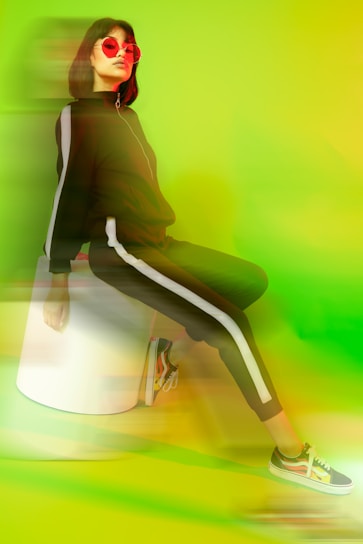 A person with short dark hair wearing a black tracksuit with white stripes, sitting on a white stool. They have on bold red sunglasses, and colorful sneakers with a vibrant lime green and yellow background creating a modern, energetic atmosphere.