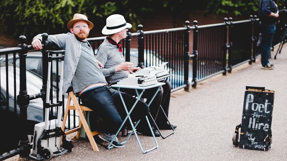 two men sitting on bench while leaning on metal rail near poet hire signboard during daytime