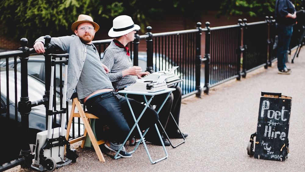 Zwei Männer sitzen tagsüber auf einer Bank, während sie sich in der Nähe des Schilds "Poet Hire" an ein Metallgeländer lehnen