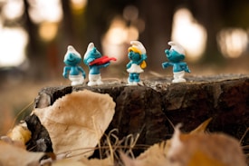 Four small blue figures with white clothing stand on a tree stump surrounded by autumn leaves. One of the figures is holding a red musical instrument, and another has yellow hair.