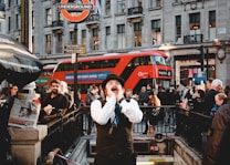 A bustling urban scene with a man in a vest and hat passionately shouting or singing in front of a busy underground station entrance. In the background, a red double-decker bus moves past, and numerous pedestrians are visible, some looking towards the man and others engaged in their activities. A street vendor's stall selling newspapers is on the left, and nearby buildings with shops are visible in the background.