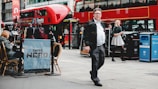 A busy city street scene with a red double-decker bus in the background and people walking and sitting at a cafe. The image features a man in business attire walking on the sidewalk and a woman using her phone. There are outdoor tables with people seated, and recycling bins are visible.