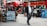 A busy city street scene with a red double-decker bus in the background and people walking and sitting at a cafe. The image features a man in business attire walking on the sidewalk and a woman using her phone. There are outdoor tables with people seated, and recycling bins are visible.