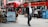 A busy city street scene with a red double-decker bus in the background and people walking and sitting at a cafe. The image features a man in business attire walking on the sidewalk and a woman using her phone. There are outdoor tables with people seated, and recycling bins are visible.