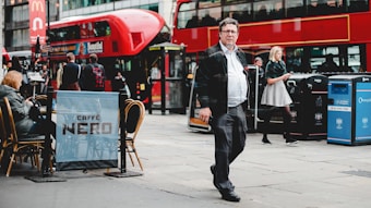A busy city street scene with a red double-decker bus in the background and people walking and sitting at a cafe. The image features a man in business attire walking on the sidewalk and a woman using her phone. There are outdoor tables with people seated, and recycling bins are visible.