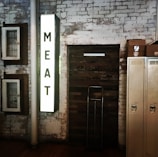A vintage industrial setting is depicted with a vertical illuminated sign displaying the word 'MEAT' on an exposed white brick wall. The background includes rustic wooden elements, old metal lockers, and stacked cardboard boxes. Nearby is a metal trolley against the wall.