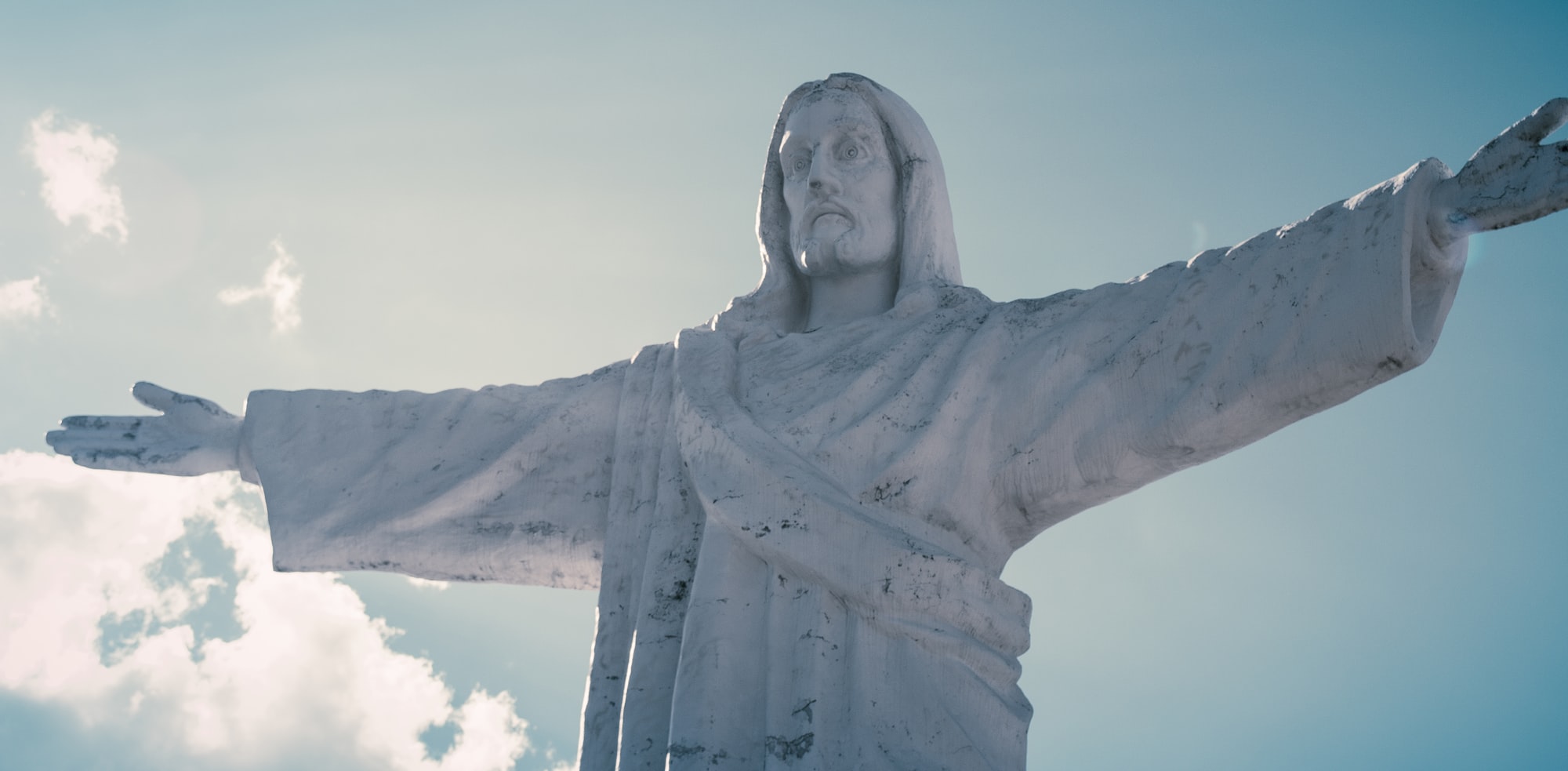 A light-colored stone sculpture of Jesus Christ with arms outstretched, blue sky and white clouds in the background.