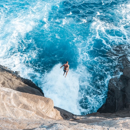 A person is captured mid-air, jumping off a rocky cliff into the vibrant blue ocean below. The waves crash against the rocks, creating a sense of motion and energy.