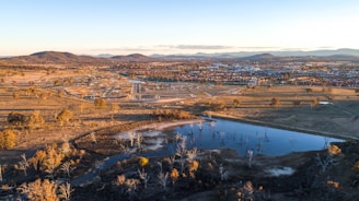 Aerial view of a semi-rural landscape featuring a body of water surrounded by dry vegetation and trees. In the background, a residential area is visible with scattered houses and buildings. Mountain ranges can be seen in the distance under a clear sky. The landscape has a mixture of natural and developed areas with roads connecting various sections.