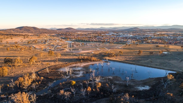 Aerial view of a semi-rural landscape featuring a body of water surrounded by dry vegetation and trees. In the background, a residential area is visible with scattered houses and buildings. Mountain ranges can be seen in the distance under a clear sky. The landscape has a mixture of natural and developed areas with roads connecting various sections.