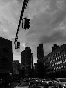A monochrome cityscape featuring tall skyscrapers silhouetted against a cloudy sky, a traffic light with signals, and cars on the street below. The scene captures the bustling energy of urban life with a mix of pedestrians and vehicles.