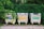 Three recycling bins with signs in a non-English language stand on a paved sidewalk, surrounded by lush green foliage. Each bin has distinct labels: blue, yellow, and green, likely indicating they are for different types of recyclables.