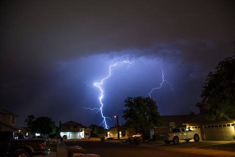 A lightning bolt illuminates the night sky over a suburban neighborhood. The street is lined with houses and trees, with parked vehicles visible. Dark storm clouds contrast against the bright flash of lightning.