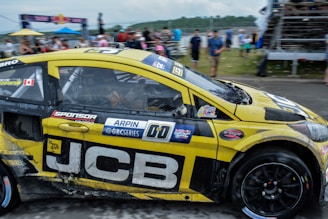 A yellow and black rally car adorned with various logos and decals races by, with its tires gripping the asphalt. Spectators can be seen in the blurred background, indicating a lively race event. The car’s exterior shows signs of dirt and wear from the track, and there is a Canadian flag sticker near the driver’s window.