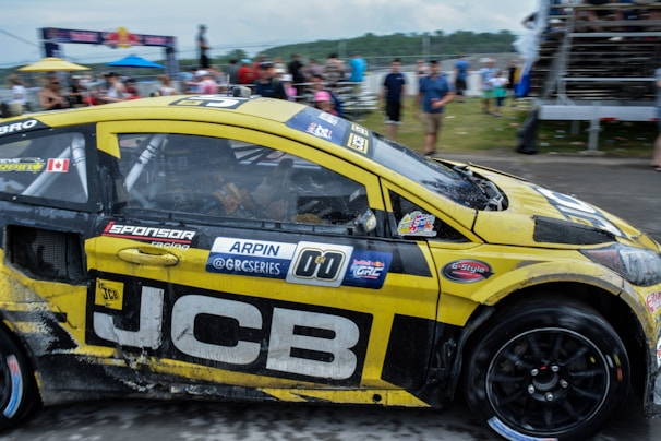 A yellow and black rally car adorned with various logos and decals races by, with its tires gripping the asphalt. Spectators can be seen in the blurred background, indicating a lively race event. The car’s exterior shows signs of dirt and wear from the track, and there is a Canadian flag sticker near the driver’s window.