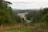 A view of a road under construction cutting through a lush green forest with some vehicles visible on it. The scene is surrounded by dense trees and vegetation, and the sky appears overcast with hills visible in the distance.