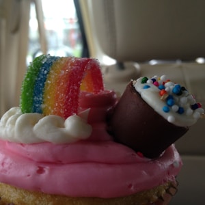 A cupcake with pink frosting topped with a candy rainbow and a chocolate piece with colorful sprinkles. The frosting is swirled and the candy creates a cheerful atmosphere.
