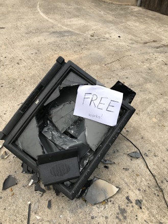 A broken television is lying on a concrete surface with shattered glass scattered around. A paper sign placed on top reads 'FREE works!'. The setting appears to be outdoors, possibly in an alley or driveway.