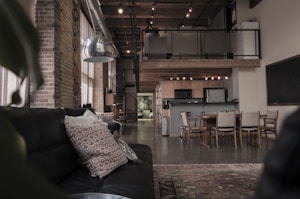 A modern, industrial-style loft with exposed brick walls and an open floor plan. The living area features a black leather couch adorned with patterned pillows. A large, metallic pendant light hangs from the ceiling. The dining area, with wooden furniture, is towards the back, near a kitchen that includes modern appliances. A spiral staircase leads to an upper mezzanine.
