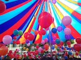 A vibrant array of colorful balloons is gathered under a large, multi-colored canopy. The balloons come in shades of red, pink, blue, purple, and green, floating upwards and filling the space under the tent. The canopy itself features bold stripes of red, blue, and yellow, creating a festive and lively atmosphere.