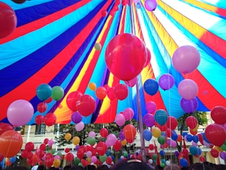 A vibrant array of colorful balloons is gathered under a large, multi-colored canopy. The balloons come in shades of red, pink, blue, purple, and green, floating upwards and filling the space under the tent. The canopy itself features bold stripes of red, blue, and yellow, creating a festive and lively atmosphere.
