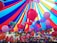 A vibrant array of colorful balloons is gathered under a large, multi-colored canopy. The balloons come in shades of red, pink, blue, purple, and green, floating upwards and filling the space under the tent. The canopy itself features bold stripes of red, blue, and yellow, creating a festive and lively atmosphere.