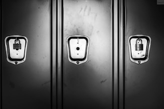 Three metal lockers are lined up side by side with a smooth, reflective surface. Two of the lockers have padlocks securing them, while the middle locker has an empty lock slot. The image is in black and white, highlighting the contrast between the shiny locks and the matte finish of the lockers.