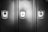 Three metal lockers are lined up side by side with a smooth, reflective surface. Two of the lockers have padlocks securing them, while the middle locker has an empty lock slot. The image is in black and white, highlighting the contrast between the shiny locks and the matte finish of the lockers.