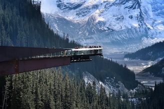 A glass-floored observation platform extends over a deep forested valley with towering snow-covered mountains in the background. Several people are standing on the platform, taking in the breathtaking views. The scene conveys a sense of adventure and natural beauty.