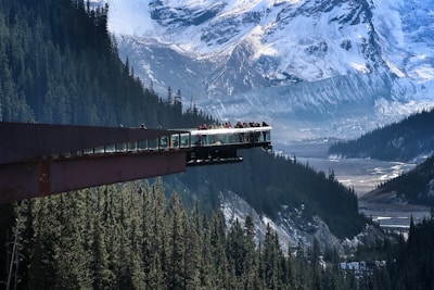 A glass-floored observation platform extends over a deep forested valley with towering snow-covered mountains in the background. Several people are standing on the platform, taking in the breathtaking views. The scene conveys a sense of adventure and natural beauty.