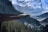 A glass-floored observation platform extends over a deep forested valley with towering snow-covered mountains in the background. Several people are standing on the platform, taking in the breathtaking views. The scene conveys a sense of adventure and natural beauty.