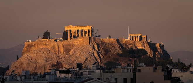A majestic ancient temple complex sits atop a rocky hill, bathed in warm, golden light during either sunrise or sunset. The surrounding modern cityscape contrasts with the historical architecture. The horizon shows distant mountains under a clear sky.
