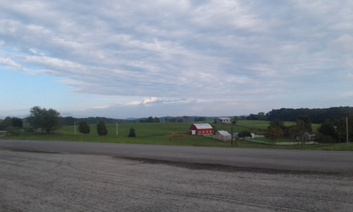 A rural landscape featuring an open expanse of green fields under a partly cloudy sky. In the distance, there is a red barn with a white roof, surrounded by various trees and other farm structures. A paved road runs across the foreground, separating the viewer from the greenery.