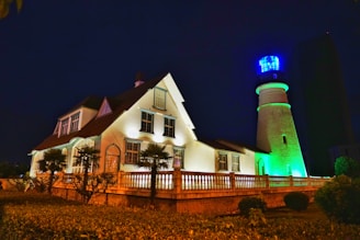A lighthouse and an adjacent house are illuminated against a dark night sky. The lighthouse emits a bright blue light at the top, while the building is lit with warm lights, creating a contrast with the night. The surrounding area includes palm trees and trimmed bushes, adding to the serene atmosphere.