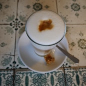 A glass of frothy coffee with a distinct espresso shot mark on top. It is placed on a white saucer with a small piece of biscotti and a silver knife beside it. The background features intricate, patterned tiles with floral and geometric designs.