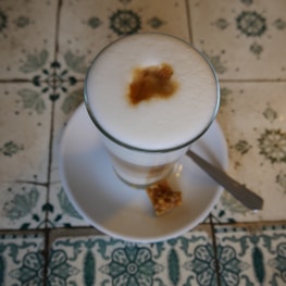 A glass of frothy coffee with a distinct espresso shot mark on top. It is placed on a white saucer with a small piece of biscotti and a silver knife beside it. The background features intricate, patterned tiles with floral and geometric designs.
