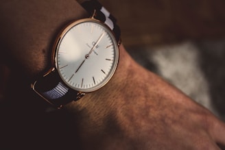 A close-up view of a minimalist wristwatch with a white face and black and white striped band. It features a rose gold case and simple hour markers with minute and hour hands pointing to approximately 10:10. The watch is worn on a wrist, highlighting the texture of the skin.