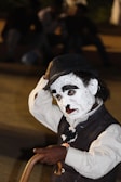 A person wearing a painted face resembling a mime or clown, with exaggerated black eyebrows and lips, dressed in a dark vest and white shirt. They are holding a cane and wearing a hat, while their expression appears thoughtful or introspective. The background is blurred, with dark tones that suggest an outdoor setting at night.