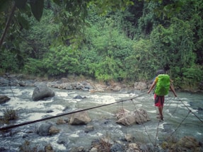 A person with a green backpack walks on a narrow makeshift bridge made of wooden planks and ropes across a fast-flowing river. The river is surrounded by lush green vegetation and large rocks, creating a sense of adventure and connection with nature.