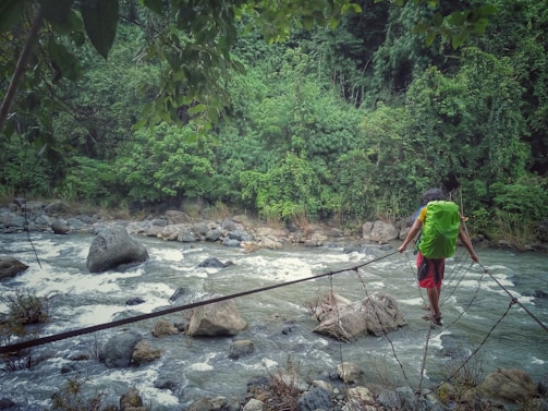 A person with a green backpack walks on a narrow makeshift bridge made of wooden planks and ropes across a fast-flowing river. The river is surrounded by lush green vegetation and large rocks, creating a sense of adventure and connection with nature.