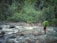 A person with a green backpack walks on a narrow makeshift bridge made of wooden planks and ropes across a fast-flowing river. The river is surrounded by lush green vegetation and large rocks, creating a sense of adventure and connection with nature.