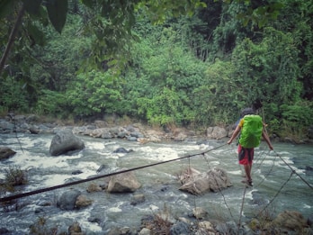 A person with a green backpack walks on a narrow makeshift bridge made of wooden planks and ropes across a fast-flowing river. The river is surrounded by lush green vegetation and large rocks, creating a sense of adventure and connection with nature.
