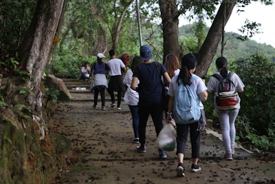 A group of people are walking along a forested path. The path is surrounded by lush greenery and large trees. Some individuals carry bags and wear casual outdoor clothing. The setting appears calm and natural, indicating a hike or outdoor walk.