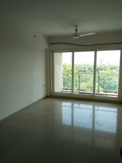 An empty, well-lit room with off-white walls and a tiled floor. There is a large window with sliding doors that open to a balcony, offering a view of a lush green landscape outside. The ceiling features a white fan situated at the center of the room.