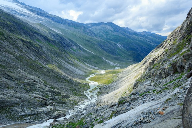 A picturesque mountain valley with a meandering river flowing through it, surrounded by steep, rocky slopes. The landscape is illuminated by diffused sunlight, revealing patches of greenery along the valley floor.