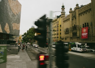 A city street featuring a blend of modern and historic architecture, including a geometric building and an ornately designed theater. A tram and several vehicles move along the street, with pedestrians walking on the sidewalks. A prominent red sign advertising a comedy festival is visible on the theater.