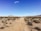 A desert landscape with a dirt path leading through sparse vegetation under a clear blue sky. In the distance, a colorful art installation featuring stacked boulders painted in bright colors stands out against the barren terrain. Mountains are visible on the horizon.