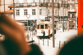 A quaint street scene features a vintage tram labeled 'Carmo' traveling along a track past historic buildings with ornate facades. Pedestrians can be seen walking nearby, and there are a variety of shop signs and traffic signals around. The overall atmosphere is bustling yet charming.
