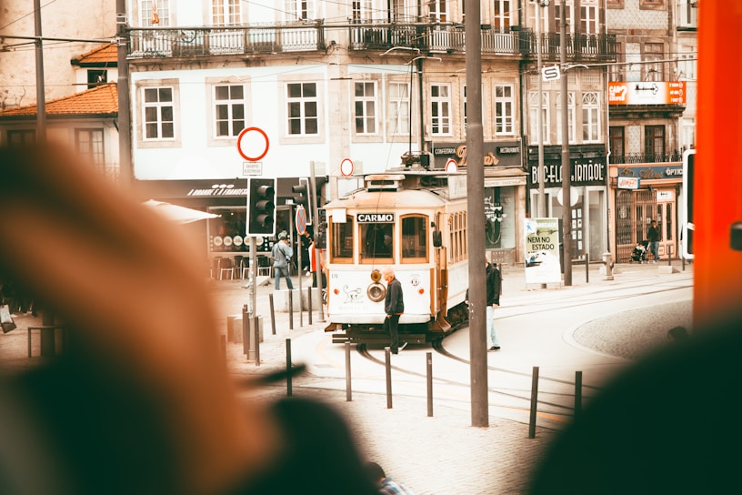 A quaint street scene features a vintage tram labeled 'Carmo' traveling along a track past historic buildings with ornate facades. Pedestrians can be seen walking nearby, and there are a variety of shop signs and traffic signals around. The overall atmosphere is bustling yet charming.