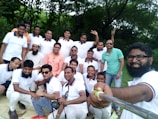A group of men gathered outdoors, some wearing matching white polo shirts with red and blue trim, with a few individuals in different shirts. One man, holding a selfie stick, takes the group photo. The background features lush green trees, suggesting a park or garden setting. The group appears cheerful and relaxed.